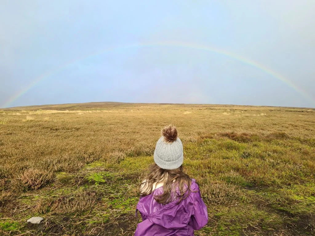 Small girl wearing a bobble hat looking at a rainbow - The Wandering Wildflower