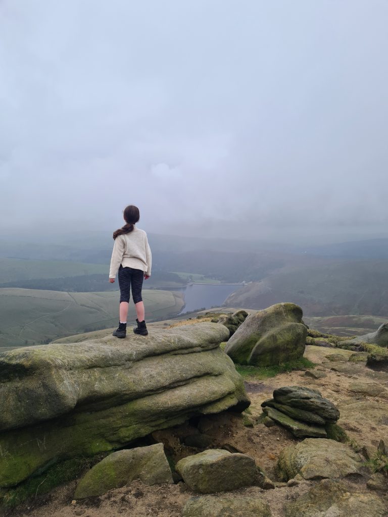 Young girl in a white fleece with black leggings on Kinder Scout, looking down to Kinder Reservoir