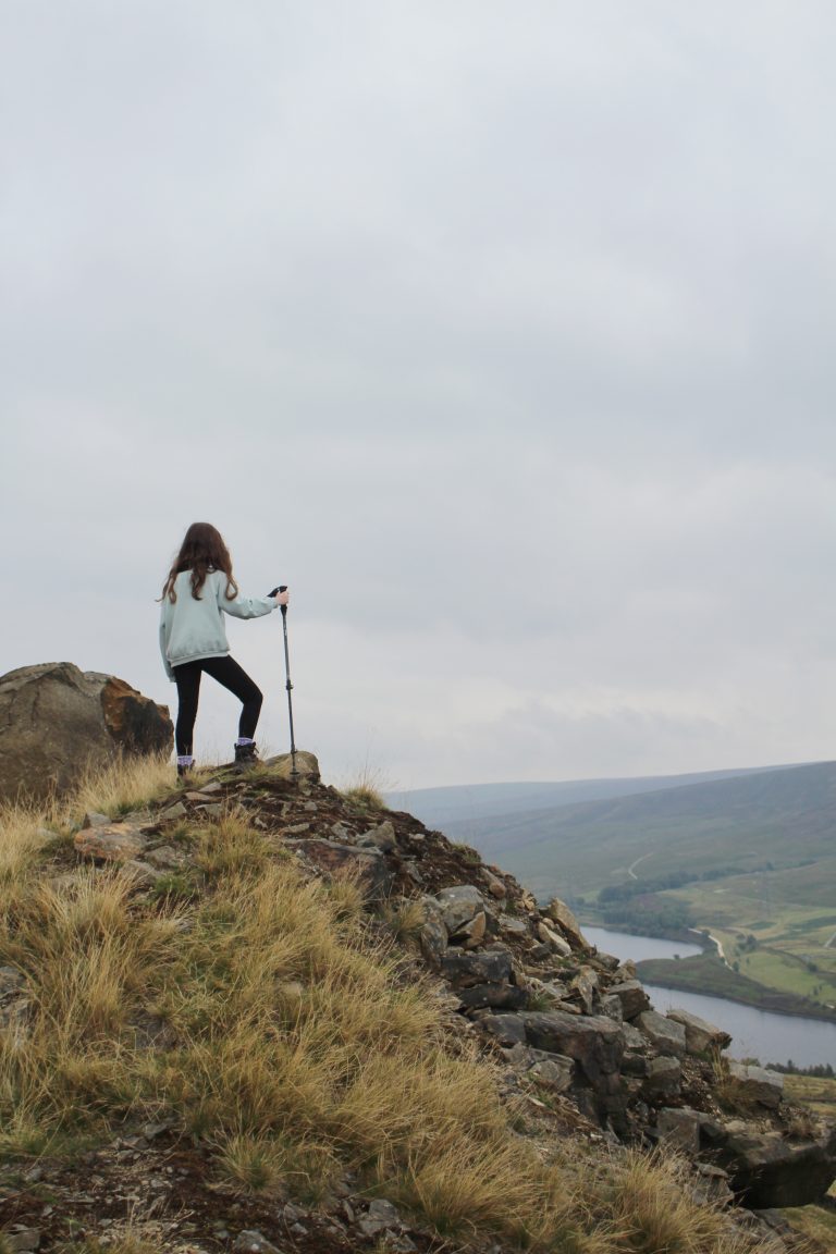 A small girl stood on a huge pile of rocks looks down to Torside Reservoir - a post on Hiking with Kids from The Wandering Wildflower