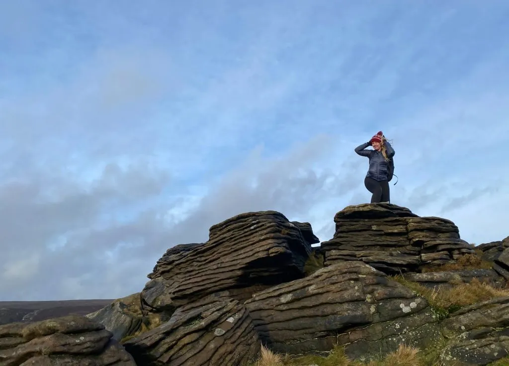 A blonde woman holds onto a bobble hat while stood on a rock formation on Grindslow Knoll