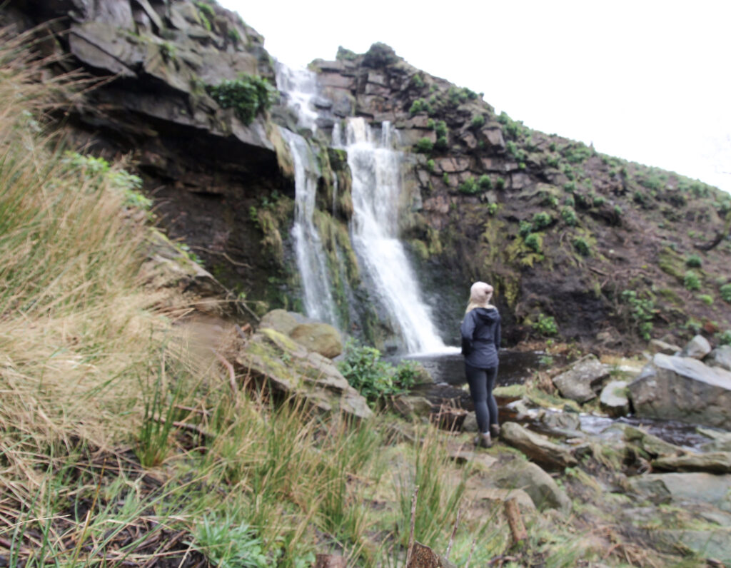 A woman dressed in black, wearing a pink bobble hat looking up at a waterfall - Wessenden waterfalls walk in the Peak District - The Wandering Wildflower