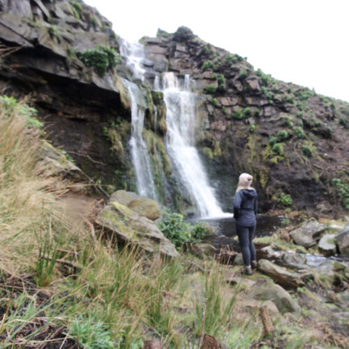 A woman dressed in black, wearing a pink bobble hat looking up at a waterfall - Wessenden waterfalls walk in the Peak District - The Wandering Wildflower