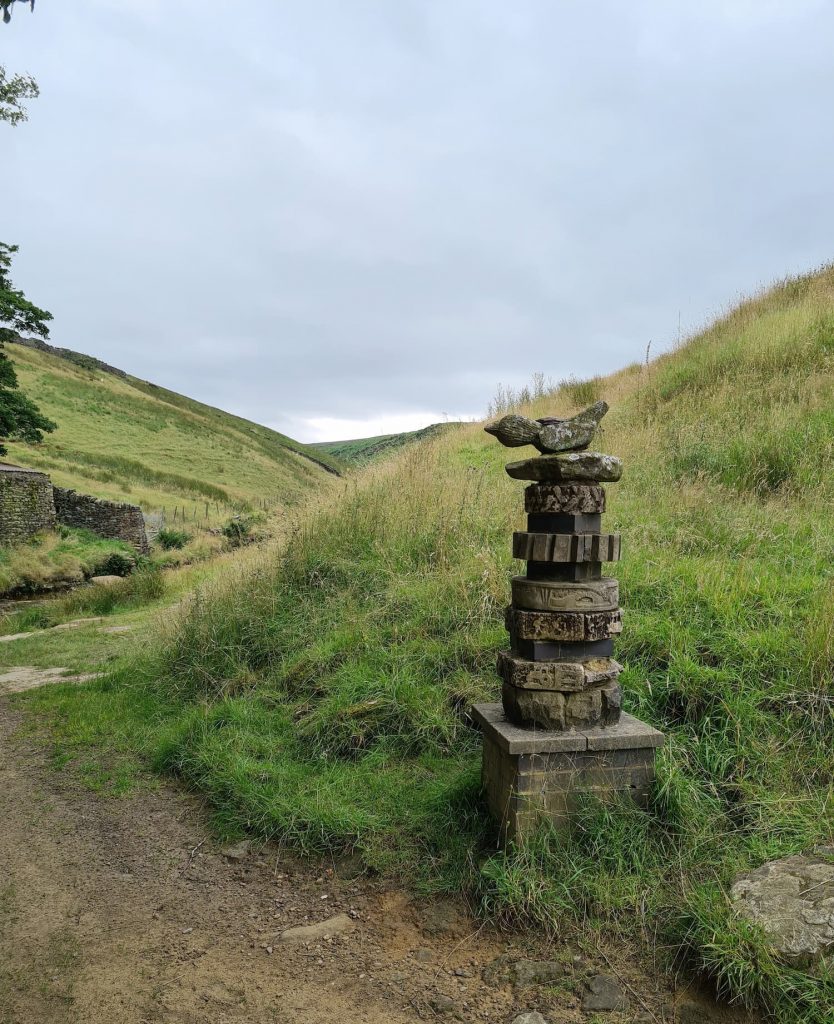 A wooden/stone sculpture with a carved bird on the top near Eastergate Bridge in Marsden, West Yorkshire