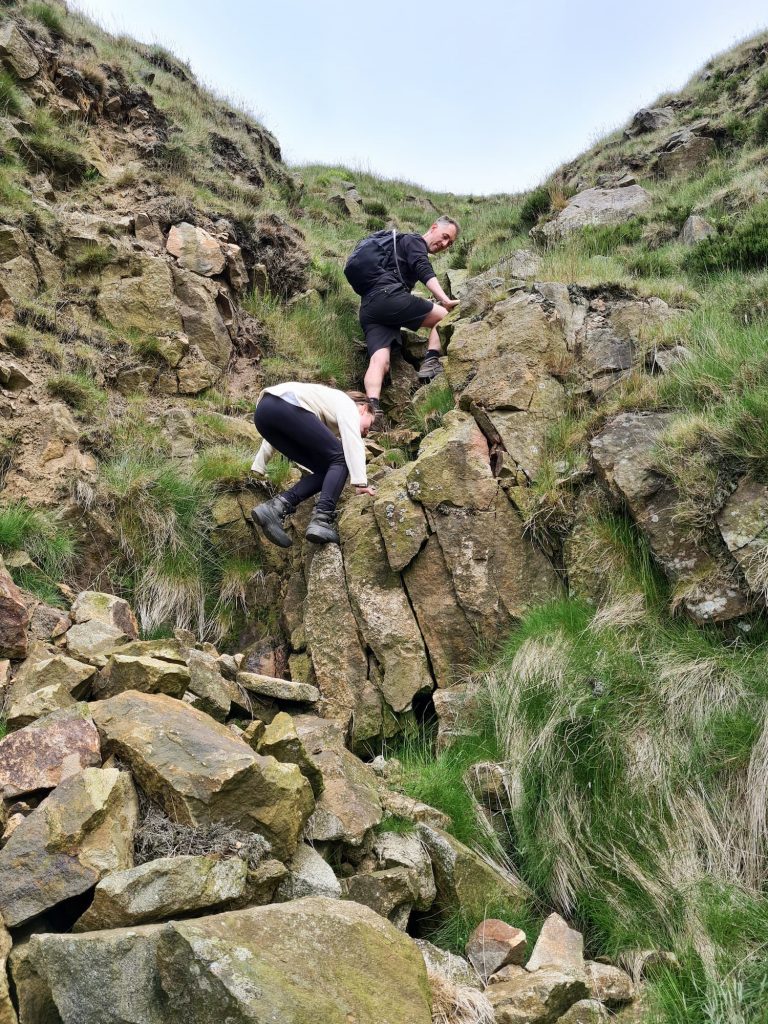 A father and daughter scrambling up a rock face in Ashton Clough, The Peak District - Higher Shelf Stones scramble walk from The Wandering Wildflower
