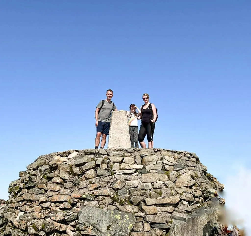 A family standing on top of the Ben Nevis summit with the trig point - Ben Nevis Route from The Wandering Wildflower