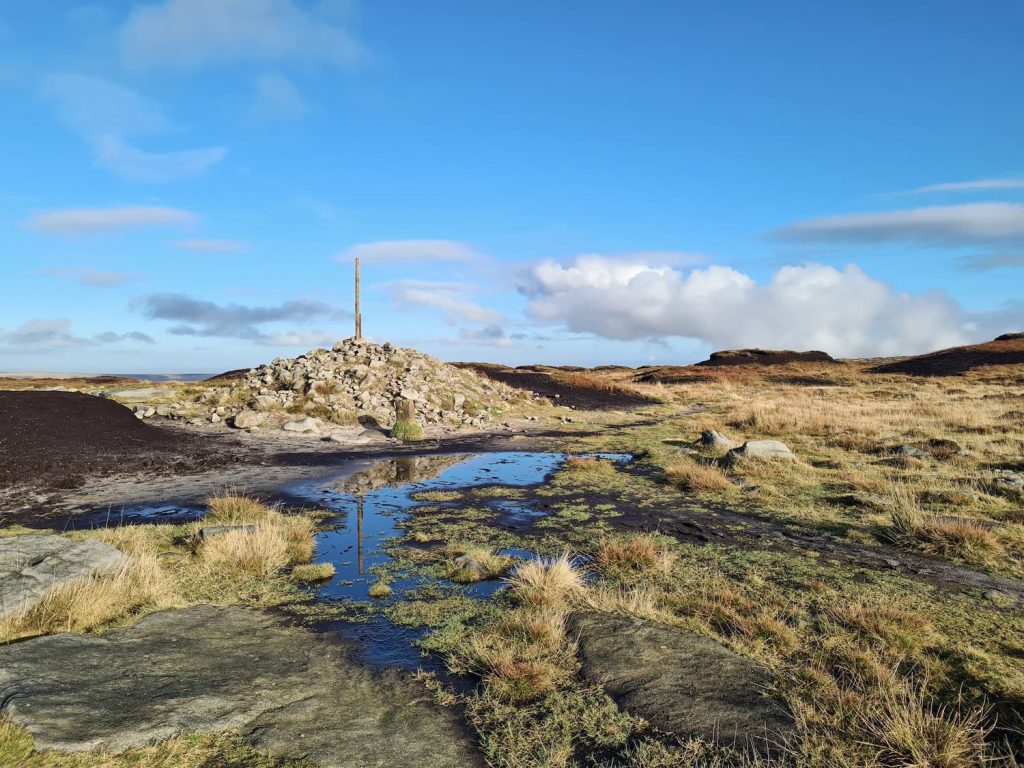 Bleaklow Head, marked with a stone cairn and a tall stake, with a reflective puddle in the foreground