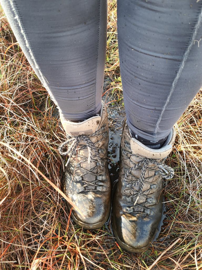 A woman's feet with brown walking boots stood in a peaty muddy puddle on the moors