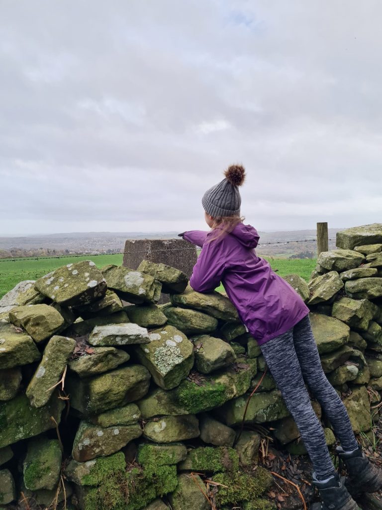 A small girl in a purple coat touching a trig point over a wall