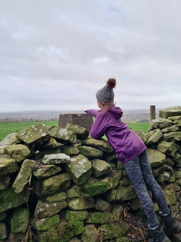 A small girl in a purple coat touching a trig point over a wall