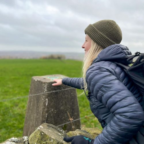 A blonde haired woman touching a trig point over a wall
