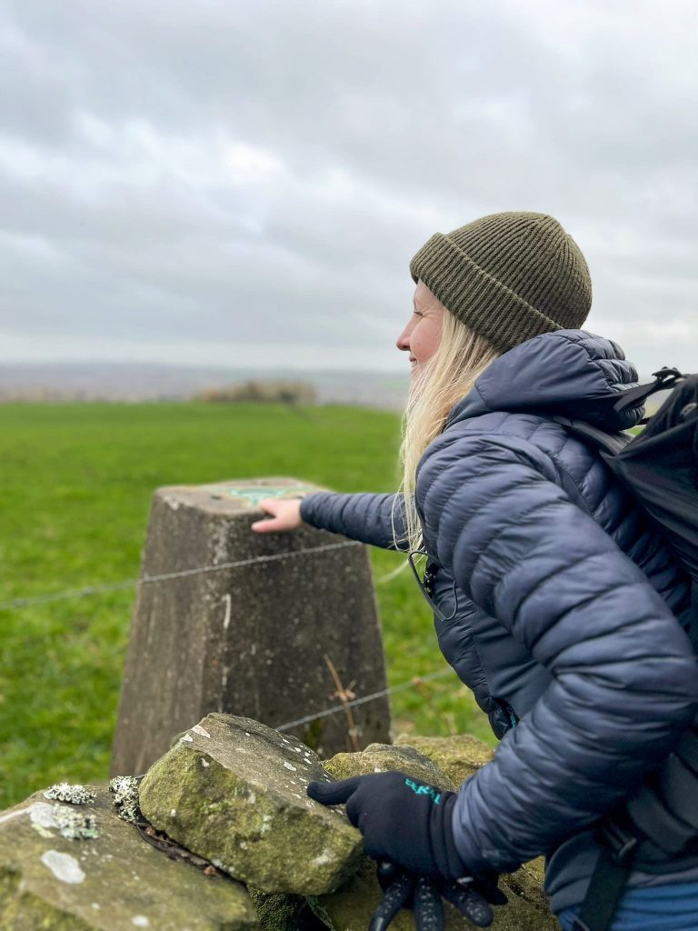 A blonde haired woman touching a trig point over a wall