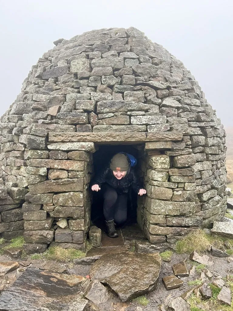 A woman in the doorway of the stone igloo on Brown Knoll. The doorway is small so she's crouching down