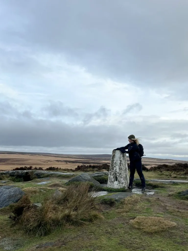 A woman standing next to White Edge trig point - it's very windy