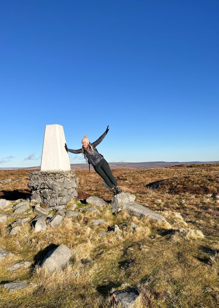 A woman standing on one trig point whilst leaning to touch another.