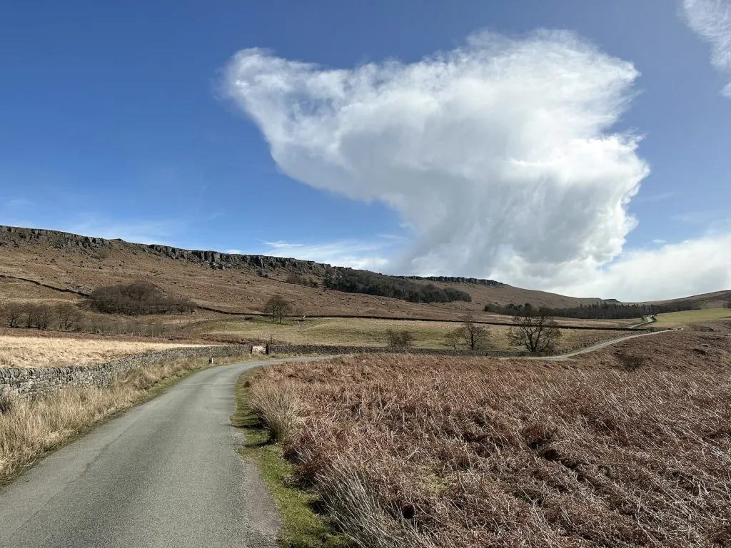 A single track road with Stanage Edge in the background