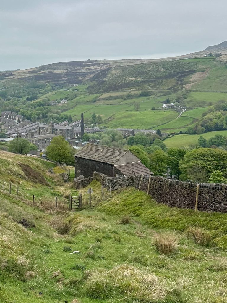 A view down into Marsden from the hillside above. There is a stone barn and a stone wall leading to a wooden gate. The hills are very green despite it being a murky day