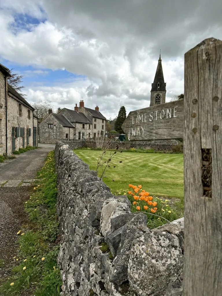 A wooden sign post for the Limestone Way pointing to a village scene, with some orange tulips