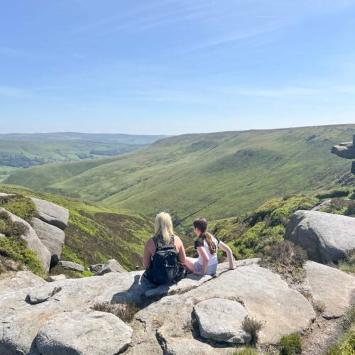 A woman and her daughter sat at the top of a hill on a rock, admiring the view of moorland and blue skies beyond