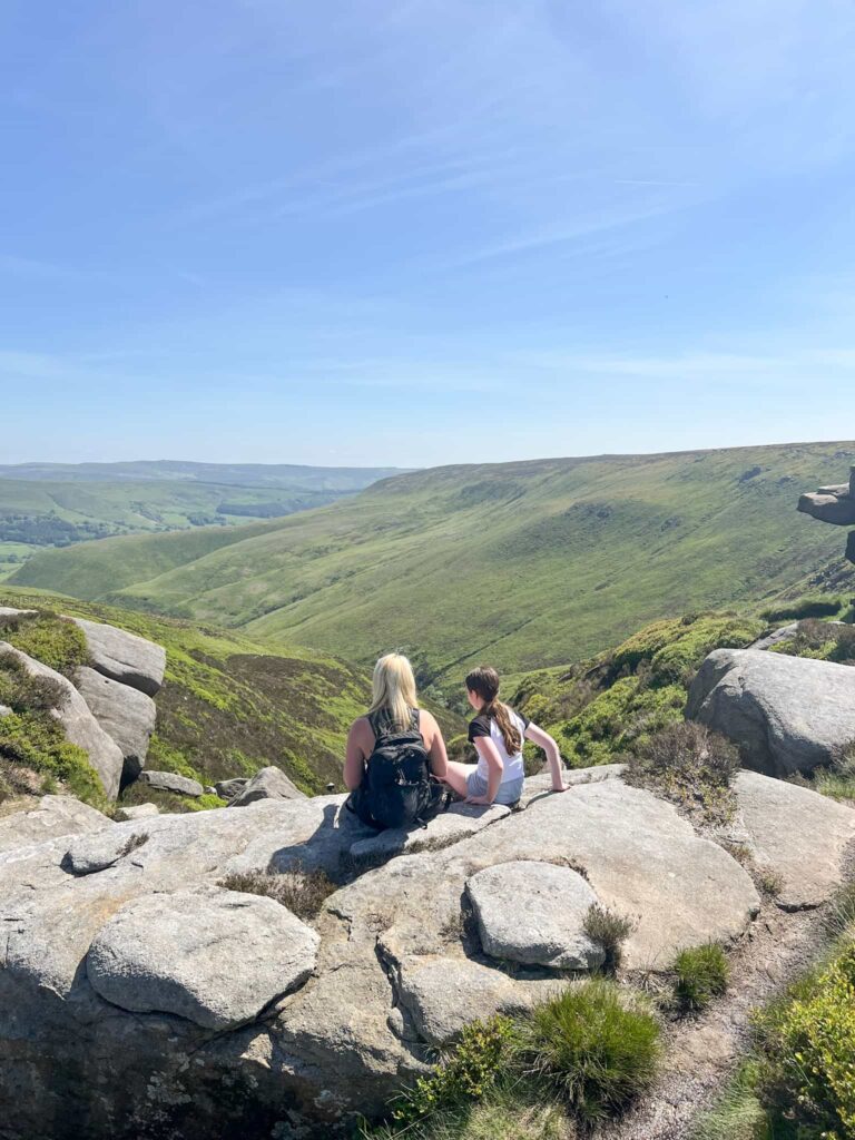 A woman and her daughter sat at the top of a hill on a rock, admiring the view of moorland and blue skies beyond