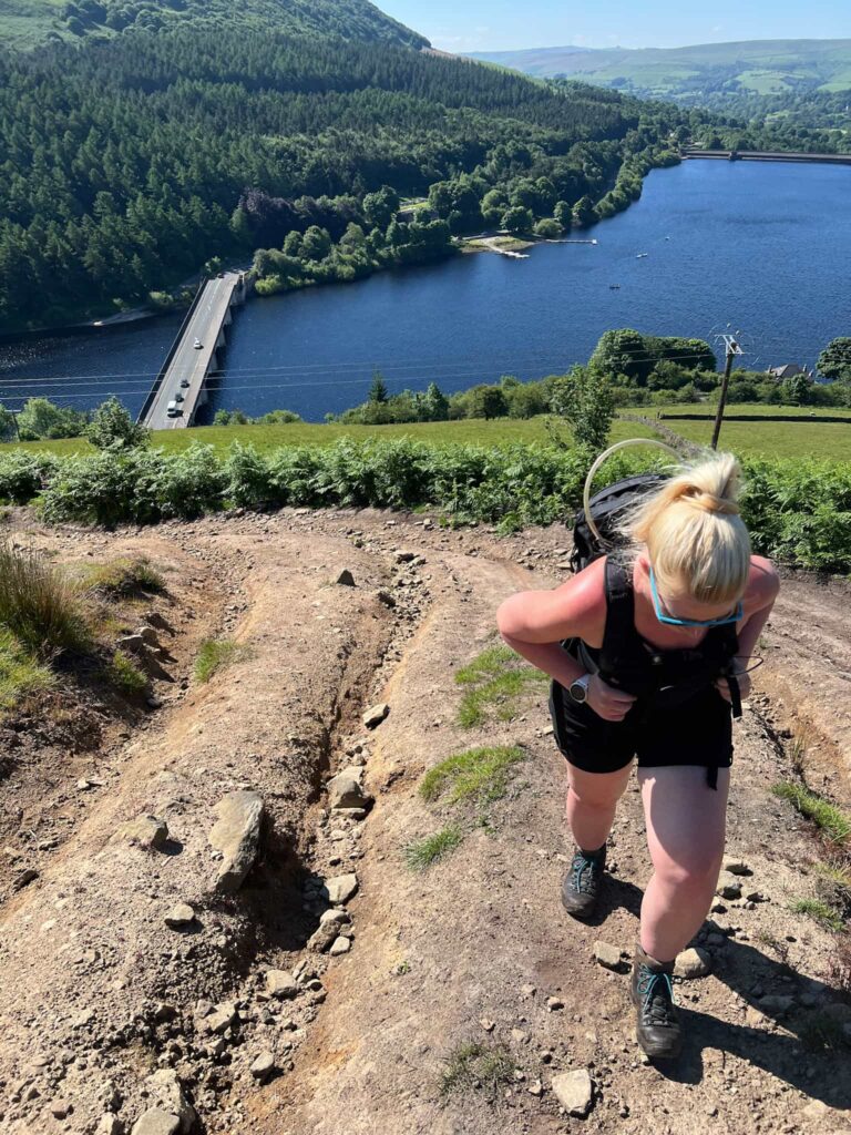 A woman with blonde hair hiking up a very steep slope, with a reservoir in the background