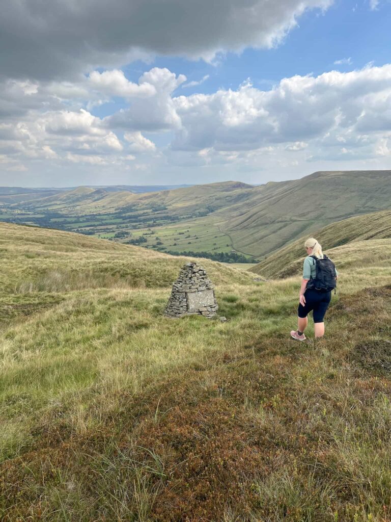A woman walking to a stone cairn with a view of the Great Ridge and Mam Tor behind
