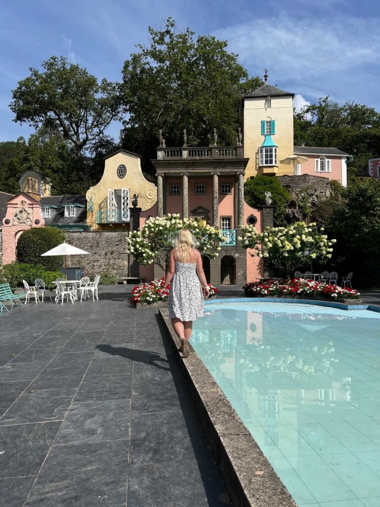A woman walking next to a clear blue pool with pastel buildings in the background. She wears a white polka dot dress and has blonde hair.