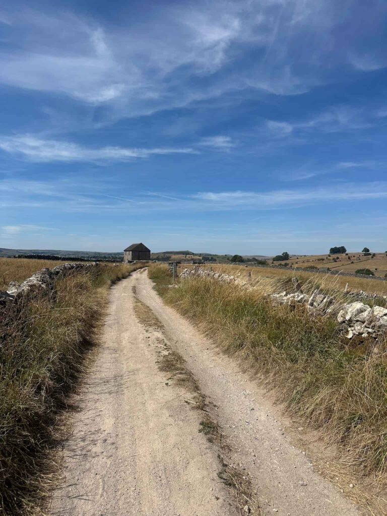 A gravel track leading into Hartington village. There are stone walls either side and a stone barn.